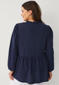 Blusa azul marino con tejido texturizado, escote con volantes y mangas largas abullonadas. Presenta un dobladillo tipo peplum y puños elásticos.