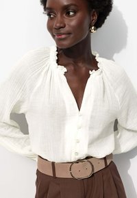 Blouse blanche texturée avec un col à volants et une fermeture à boutons, associée à un pantalon marron taille haute et une ceinture beige avec une boucle ronde.