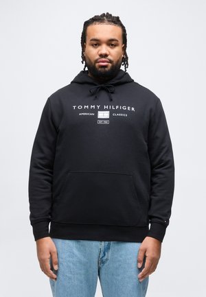 Moški s spetimi lasmi, oblečen v črno Tommy Hilfiger kapucarico in svetlo modre kavbojke, stoji pred enostavnim belim ozadjem.