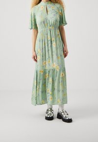 Langes, mintgrünes Kleid mit floralem Muster, kurzen Ärmeln und Schleifenkragen. Hat einen gestuften Saum und wird mit metallischen Plateauschuhen getragen.