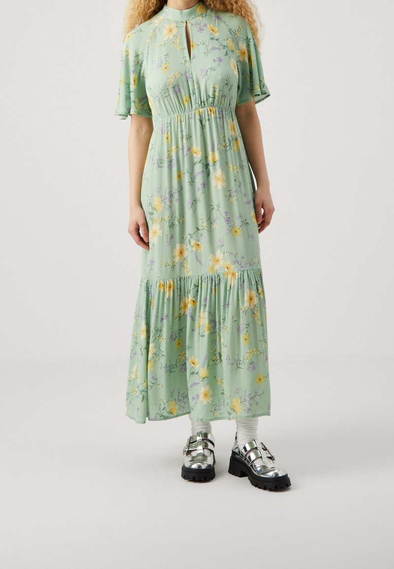 Langes, mintgrünes Kleid mit floralem Muster, kurzen Ärmeln und Schleifenkragen. Hat einen gestuften Saum und wird mit metallischen Plateauschuhen getragen.