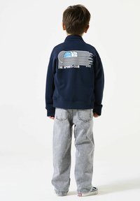 Donkerblauwe sweatshirt met hoge kraag, voorzien van een grafisch ontwerp in wit en lichtblauw, gecombineerd met lichtgrijze baggy jeans. Casual look.