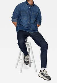 Donkerblauwe denimhemd met twee borstzakken, gecombineerd met donker slim-fit jeans en zwart-witte sport Sneakers. Witte houten kruk.