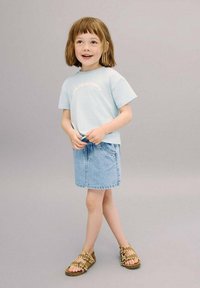 Vaaleansininen lyhythihainen T-paita, jossa on teksti "RAKASTA TUOREITA HEDELMIÄ", yhdistettynä farkkuhameeseen ja ruskeisiin sandaaleihin, joissa on pyöreitä yksityiskohtia.