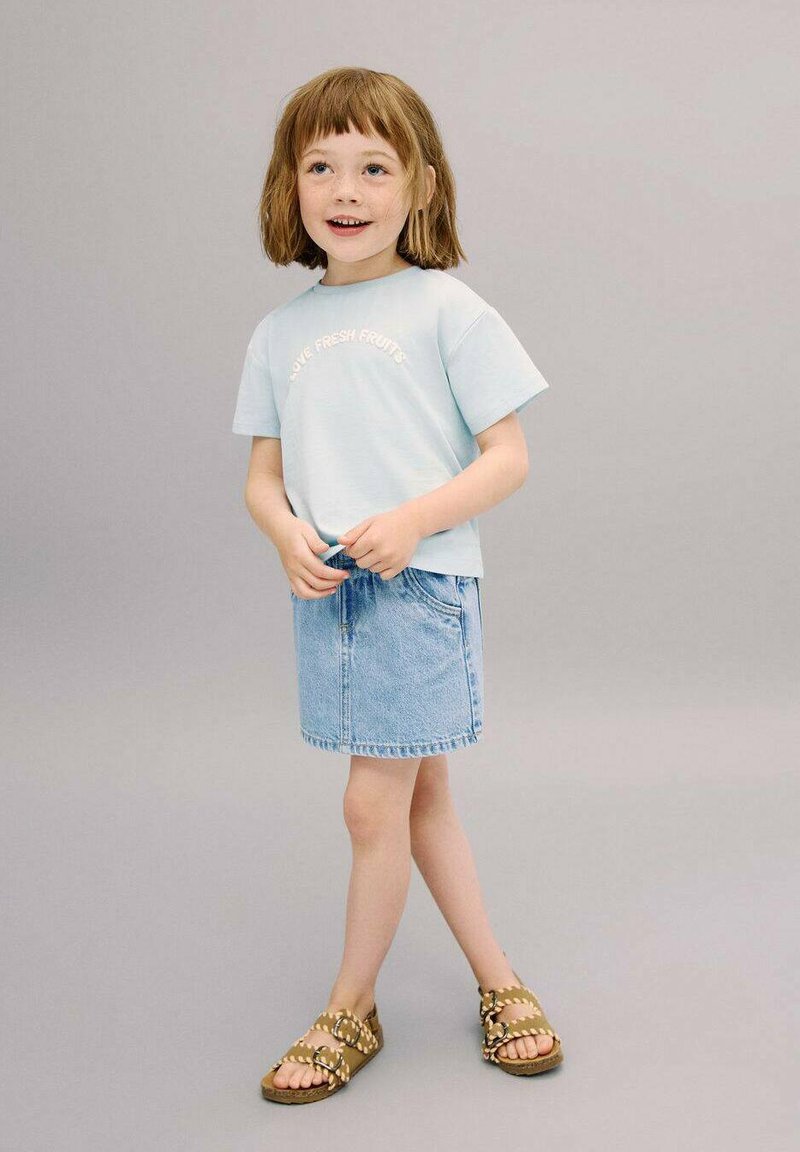 Vaaleansininen lyhythihainen T-paita, jossa on teksti "RAKASTA TUOREITA HEDELMIÄ", yhdistettynä farkkuhameeseen ja ruskeisiin sandaaleihin, joissa on pyöreitä yksityiskohtia.