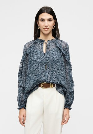 Žena nosi priehľadnú tmavomodrú kvetovanú blúzku s volánovým golierom a rukávmi, biele nohavice s vysokým pásom a hnedý opasok so zlatou prackou.