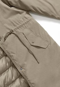 Giacca isolante beige con tessuto trapuntato, coulisse regolabile e una tasca con bottone a pressione. Tessuto liscio con hardware minimo.
