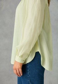 Blusa leggera gialla a maniche lunghe con una vestibilità rilassata, polsini abbottonati e orlo curvo, abbinata a jeans blu scuro. Tessuto liscio e strutturato.