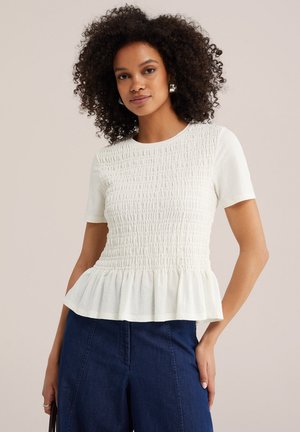 Frau mit lockigem Haar trägt ein weißes, gerafftes kurzärmliges Oberteil mit Peplum-Saum und dunkelblaue, hoch taillierte Hose, steht mit einer Hand in der Tasche.