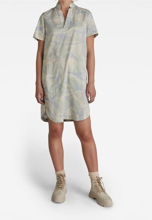 Personne portant une robe courte à manches courtes, camouflage clair, longueur genou, avec deux poches avant, chaussettes beiges et bottines beiges à lacets.