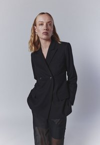 Melna dubultā jaka ar ieliektām atloks, divām priekšējām kabatām un gludu tekstūru. Uzvilkta virs caurspīdīgām melnām tīkla biksēm.