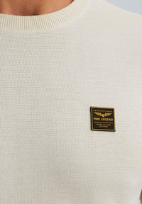 Maglione lavorato a maglia color crema con un motivo testurizzato e colletto a coste. Presenta un'etichetta con logo nero e oro sul petto.