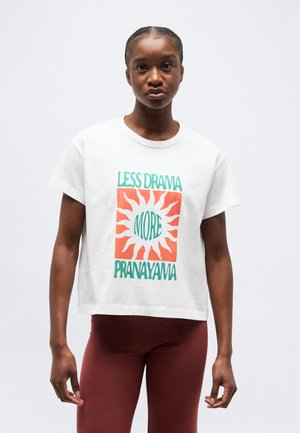 Joven mujer con camiseta blanca con gráfico de sol naranja y texto "Menos drama, más pranayama" y leggings granate, de pie contra un fondo blanco.