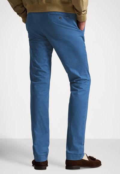 Calças de algodão azuis com corte direito, com bolsos traseiros, textura subtil e bainha dobrada. Usadas com mocassins marrons.