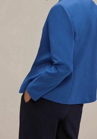 Blazer bleu cintré en tissu lisse, avec une coupe ajustée, une fermeture à un bouton, et des revers crantés élégants. Associé à un pantalon foncé.