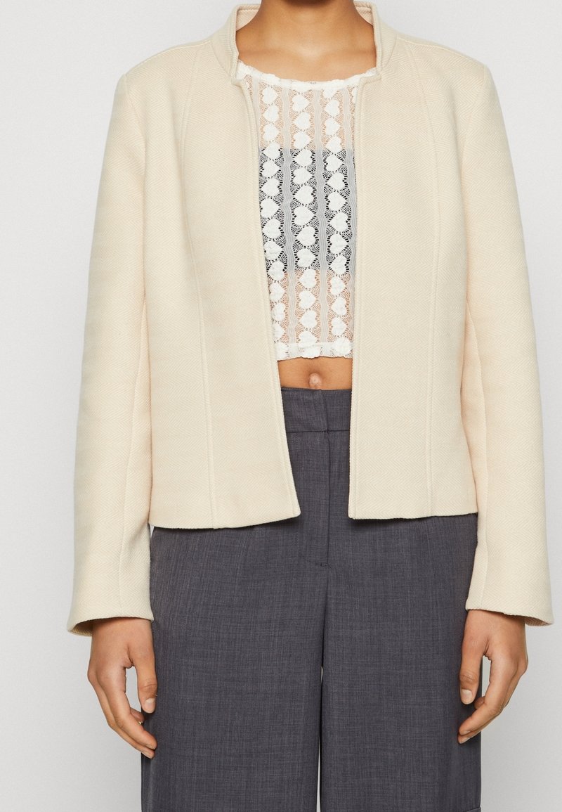Personne portant un blazer de couleur crème sur un crop top en dentelle blanche, associé à un pantalon gris taille haute et ajusté, debout devant un fond uni.