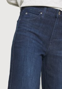 Detailní záběr na pas a horní stehna tmavě modrých džínsů s viditelnou přední kapsou, poutkem na pásek a knoflíkem.