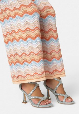Pieds portant des sandales argentées à talons hauts avec des brides ornées de strass, associées à un pantalon large présentant des rayures ondulées pastel orange, bleu et beige.