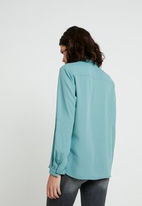 Femme aux cheveux bruns mi-longs portant un chemisier à manches longues couleur sarcelle et un jean gris foncé, vue de dos sur un fond blanc uni.