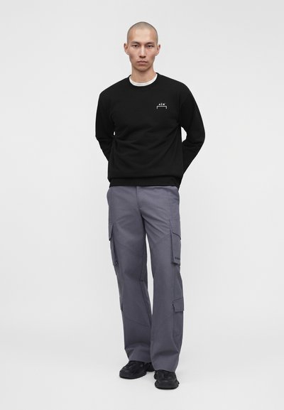 Άνδρας όρθιος, φοράει μαύρο φούτερ με μικρό λευκό λογότυπο, γκρι παντελόνι cargo και μαύρα παπούτσια, πάνω σε απλό ανοιχτόφωτο φόντο.