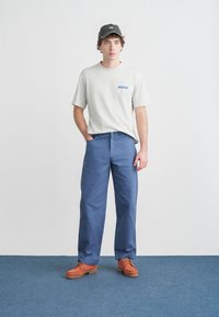 Γκρι κοντομάνικο μπλουζάκι με μπλε λογότυπο «Dickies», συνδυασμένο με μπλε παντελόνι και πορτοκαλί παπούτσια. Το μοντέλο φοράει σκουρόχρωμο καπέλο και στέκεται σε μπλε δάπεδο.