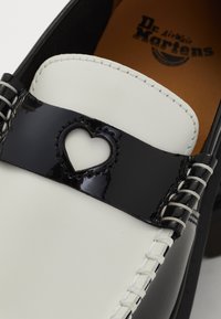 Zwart-witte schoen met een lakleren band, voorzien van een hartvormige uitsnede, contrasterende stiksels en het Dr. Martens logo op de binnenzool.