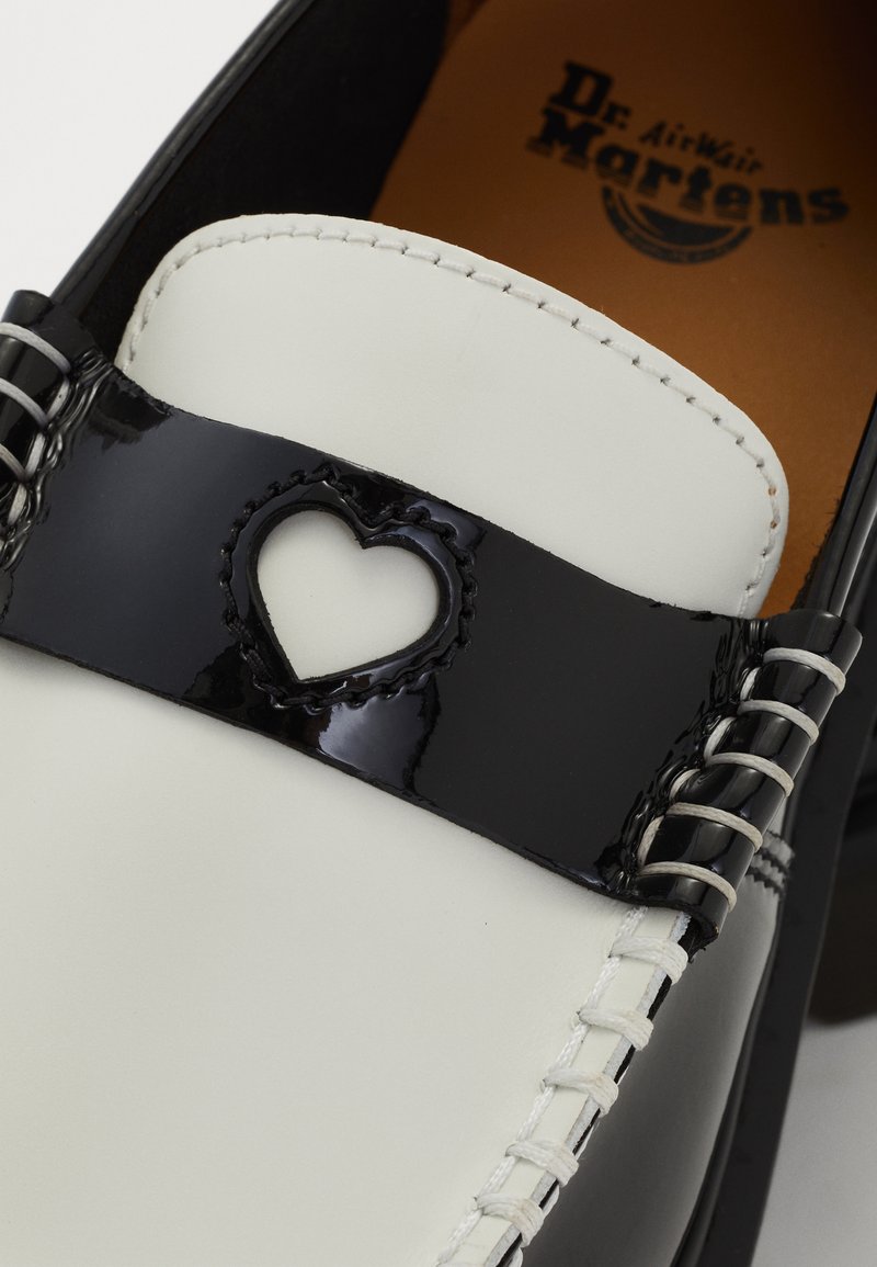 Chaussure noir et blanc avec une bande en cuir verni, présentant une découpe en forme de cœur, des coutures contrastées et le logo Dr. Martens sur la semelle intérieure.