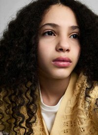 Närbild av en ung person med mörkt, lockigt hår, iklädd ett gult stickat plagg över en vit topp. Neutral bakgrund.