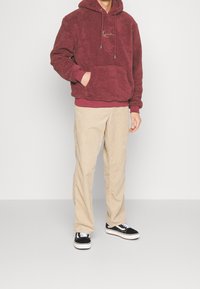 Sudadera de forro polar burdeos con bolsillo tipo canguro y cordón, combinada con pantalones de pana beige y zapatillas negras Vans.