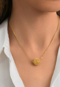 Collier pendentif en or présentant un charme sphérique texturé, suspendu à une chaîne délicate, porté sur une chemise blanche.