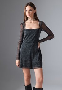 Vestido negro con brillo, mangas largas transparentes, escote cuadrado, cintura ajustada y pantalones cortos sueltos. Acentuado con un collar choker en forma de corazón.