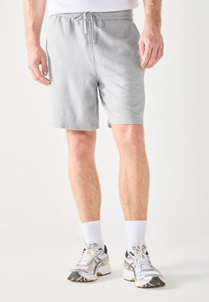 Bermudas gris con cordón y bolsillos laterales, hechas de una tela texturizada. Combinadas con calcetines deportivos blancos y zapatillas de deporte con acentos en negro y amarillo.