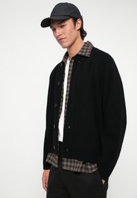 Moški nosi črno kapo, črno gumbno jopico čez rjavo karo srajco in belo majico, stoji z eno roko v žepu na belem ozadju.