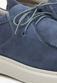 Chaussure en cuir bleu avec une finition texturée, présentant des coutures blanches et des lacets gris. La semelle est de couleur claire et légèrement contourée.