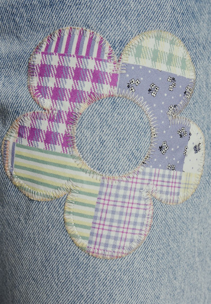 Tela de mezclilla con un parche de flores hecho de telas mixtas en patrones de cuadros púrpura, verde y azul, con un agujero central y costuras visibles.