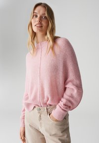 Maglione rosa chiaro lavorato a maglia con collo alto e maniche larghe. La texture è soffice, con una cucitura centrale sul davanti.