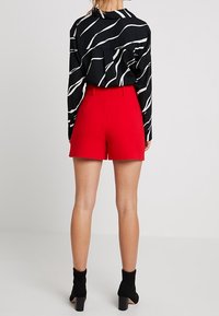 Žena oblečená v černobíle vzorované blůze s dlouhým rukávem, světlých červených šortkách s vysokým pasem a černých botách na podpatku, stojící zády.