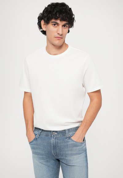Νεαρός άντρας με σγουρά μαύρα μαλλιά και σκουλαρίκια φοράει ένα απλό λευκό t-shirt και ανοιχτόχρωμα μπλουτζίν, στέκεται με τα χέρια στις τσέπες.