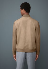 Hellbraune Bomberjacke mit einem gerippten Kragen, glatter Textur und elastischen Bündchen. Sichtbare Nähte und lockere Passform. Grauer Hintergrund.