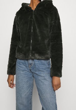 Personne portant une veste à capuche zippée pelucheuse vert foncé et un jean en denim bleu clair sur un fond uni clair.