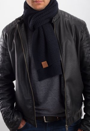 Knitted navy scarf with textured pattern, featuring a tan leather label, worn over a gray shirt and black leather jacket.