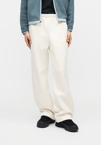 Άτομο που φοράει άνετο λευκό παντελόνι φόρμας, μαύρα αθλητικά παπούτσια, μαύρο μπλουζάκι και μπλε φλις μπουφάν με φερμουάρ, στέκεται μπροστά σε απλό φόντο.