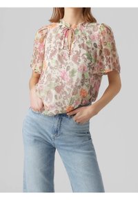 Blouse fleurie en tissu léger avec un col volanté et un détail noué, présentant des motifs roses, orange et verts. Associée à un jean bleu clair.