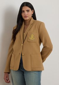 Beige blazer med teksturert stoff, enkelt knappelukking, to frontlommer og en gullbrodert logo på brystlommen.