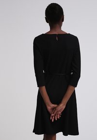 Vestido negro hasta la rodilla con mangas tres cuartos, textura acanalada y detalle de abertura en la espalda. Cintura ajustada con cinturón.