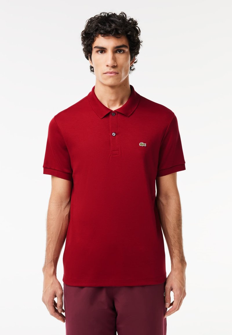 Rød bomulds polo shirt med krave, tre-knaps lukning, korte ærmer og lille grøn logo på venstre bryst.