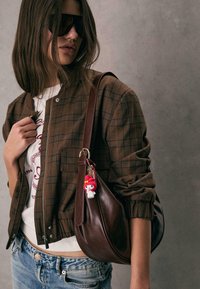 Giacca bomber a scacchi marroni, t-shirt grafica bianca, pantaloni shorts in denim e borsa a spalla in pelle marrone con un portachiavi di un personaggio come dettaglio.