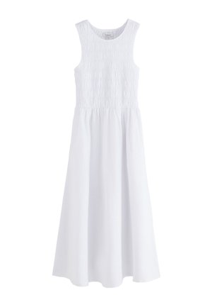 Donna che indossa un vestito midi bianco senza maniche con schiena arricciata, in piedi vicino a una moderna scala a chiocciola in legno all’interno.