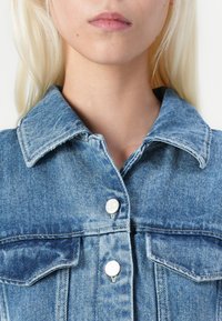 Giacca di jeans azzurra chiara con colletto a punta, due tasche sul petto, bottoni di metallo argentato e una finitura in tessuto testurizzato.