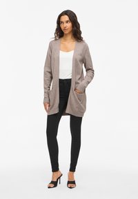 Lång, öppen cardigan i ljusbrunt stickat material, med två framfickor och rippade muddar, som är kombinerad med svarta slim-fit byxor och klackar.